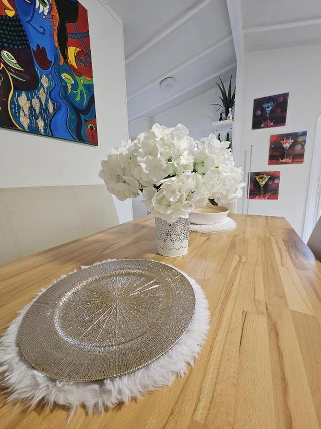 Een eettafel met een geweven placemat, een vaas met witte bloemen en een kleurrijk schilderij aan de muur.