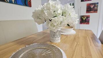 Een eettafel met een geweven placemat, een vaas met witte bloemen en een kleurrijk schilderij aan de muur.