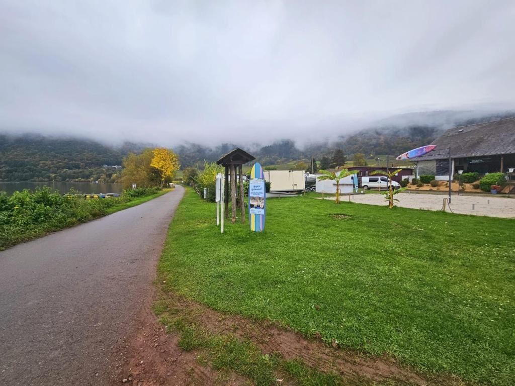 Ein gepflasterter Weg führt zu einem Gebäude mit einer kleinen Schutzhütte, einem Schild und einer Rasenfläche, im Hintergrund ist ein Berg zu sehen.
