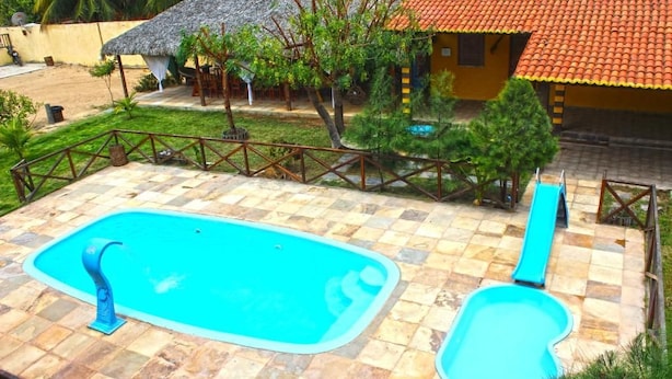 Una piscina con tobogán, rodeada por una zona vallada con una estructura de techo de paja y una casa con techo de tejas.