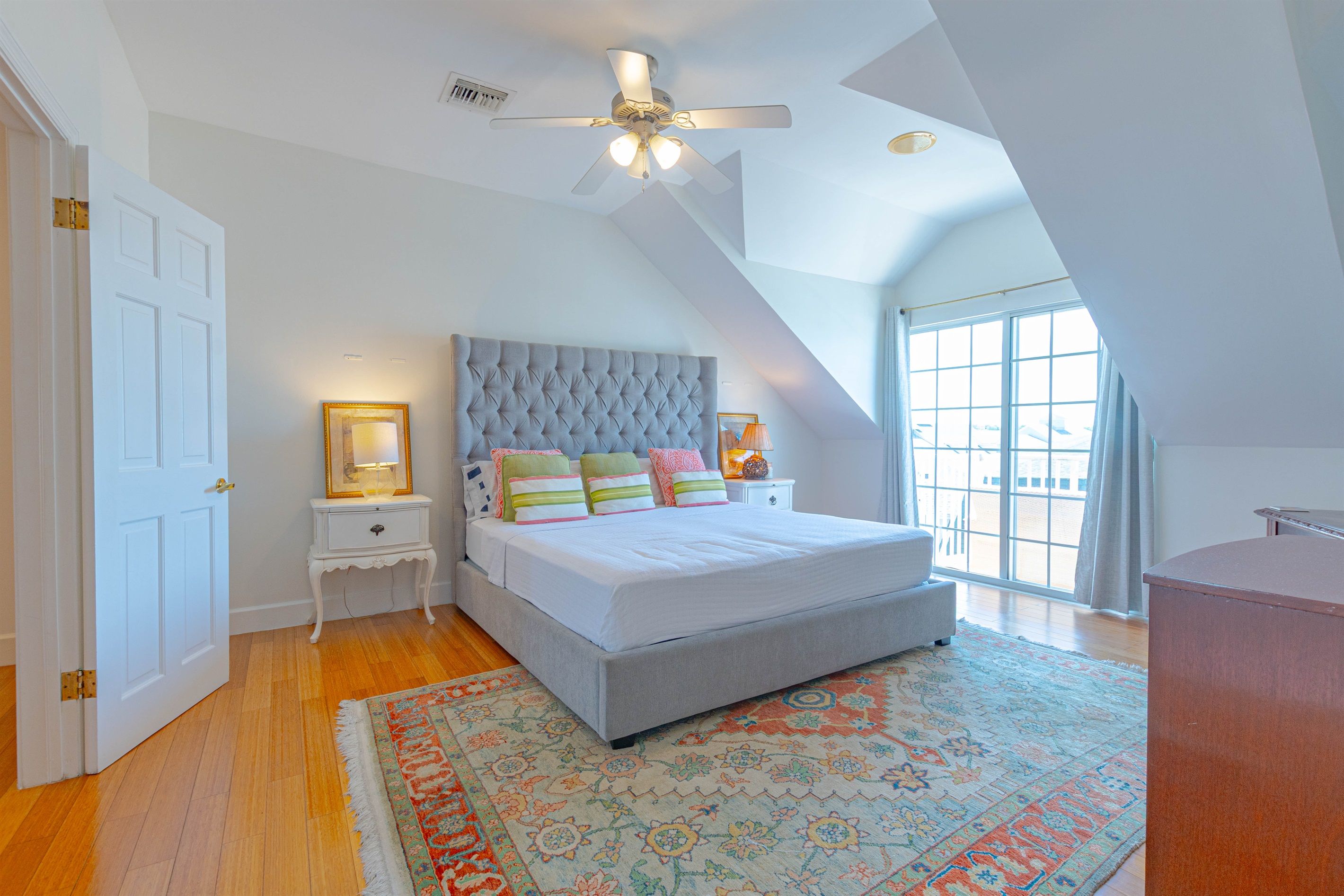 A bedroom with a large bed, a ceiling fan, a nightstand, a window with curtains, and a patterned rug.