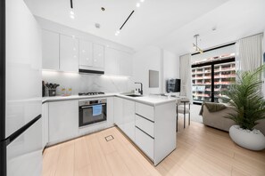 A modern kitchen with white cabinetry, a built-in oven, and a dining area with a view of a construction site. - Stayshort - Peace and Tranquility in This 1 Bedroom in JVC (Dubai)