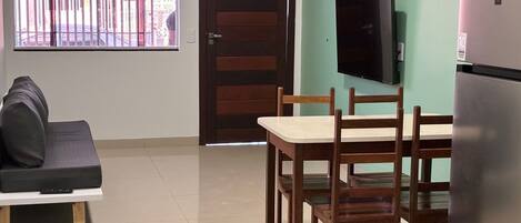 A compact living space with a sofa, a dining table with chairs, a TV mounted on the wall, and a refrigerator.