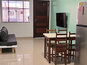A compact living space with a sofa, a dining table with chairs, a TV mounted on the wall, and a refrigerator.
