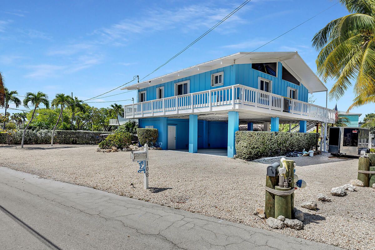 A two-story blue house with a balcony, surrounded by gravel and palm trees.