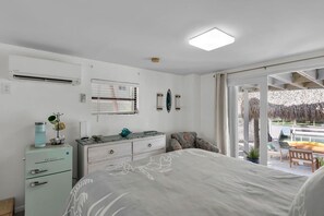 Ein Schlafzimmer mit Bett, Kommode, Fenster mit Jalousien, Deckenventilator und Blick auf einen Außenbereich mit Tisch und Stühlen.