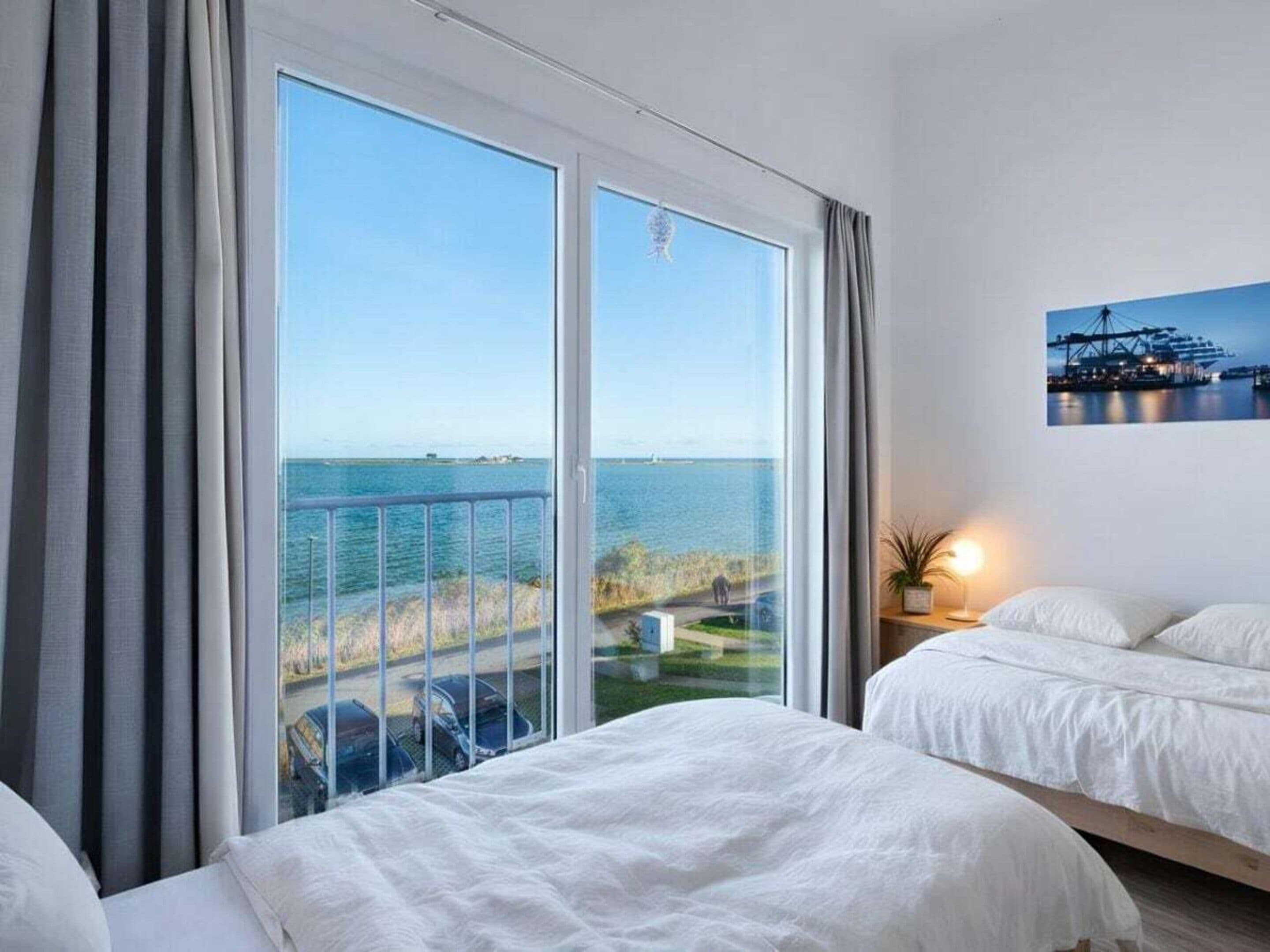 A hotel room with a large window offering a view of the sea and a distant island. There are two beds with white linens and a small bedside table with a lamp.