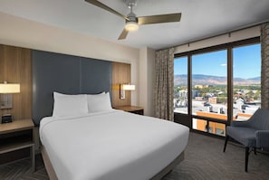 A hotel room with a large bed, a ceiling fan, a view of a cityscape through large windows, and a chair. - BLOCK Blue Ridge Village 2br (Banner Elk)