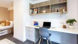 A modern bedroom with a white bed, a desk with a laptop, a chair, and a shelf with decorative items. - Student Only The Gillows (Lancaster)