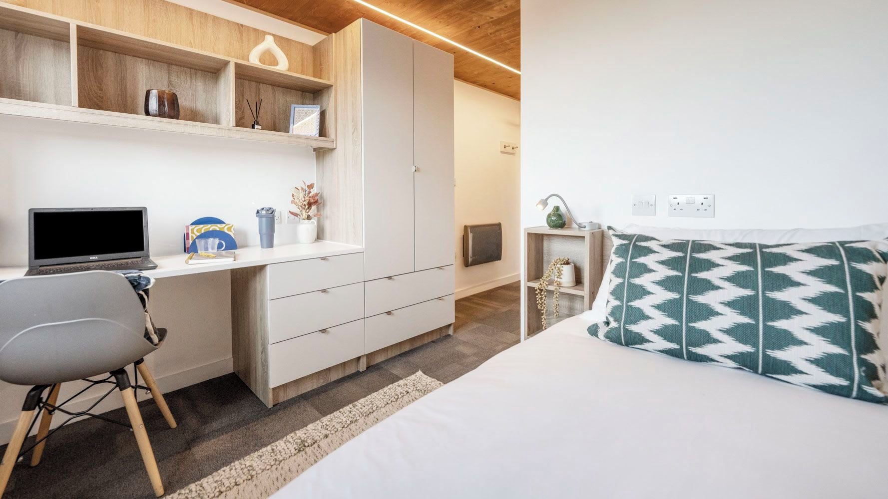 A bedroom with a bed, a desk with a laptop, a chair, and a shelf with decorative items.