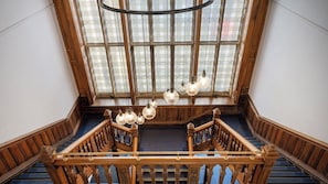 A grand staircase with wooden railings and a blue carpet, leading to a room with large windows and a chandelier. - Student Only The Gillows (Lancaster)