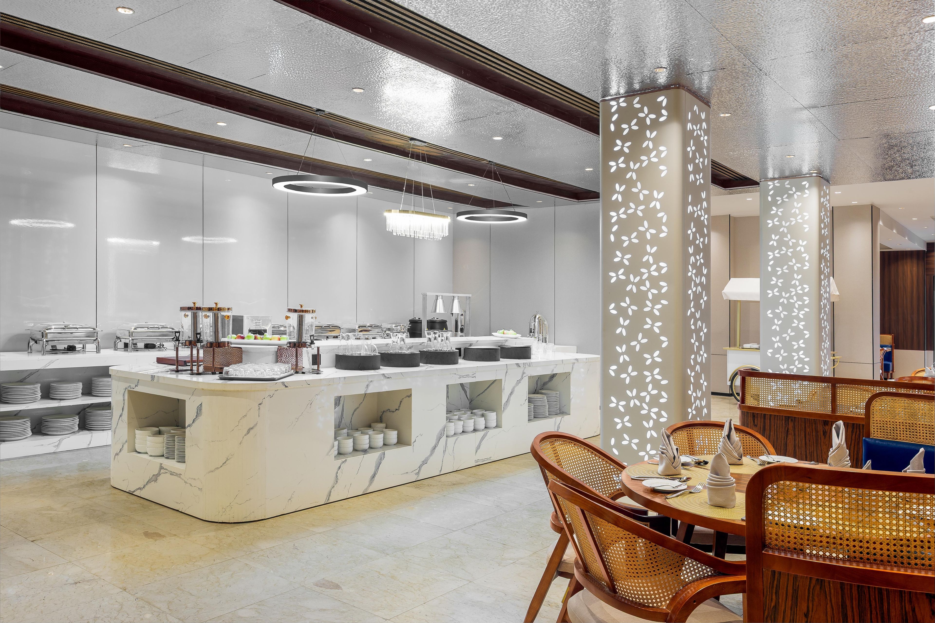 Un hall d’hôtel moderne avec un comptoir d’accueil en marbre, un espace repas doté de chaises en osier et un plafond pourvu d’un éclairage encastré.
