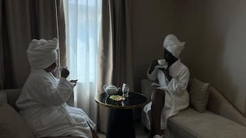 Two individuals in white bathrobes sitting on a sofa and a small round table with a teapot and a cup between them.