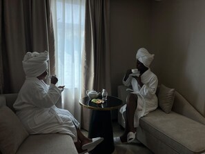 Two individuals in white bathrobes sitting on a sofa and a small round table with a teapot and a cup between them. - Best Western Plus Ambience Hotel Ikeja (Lagos)