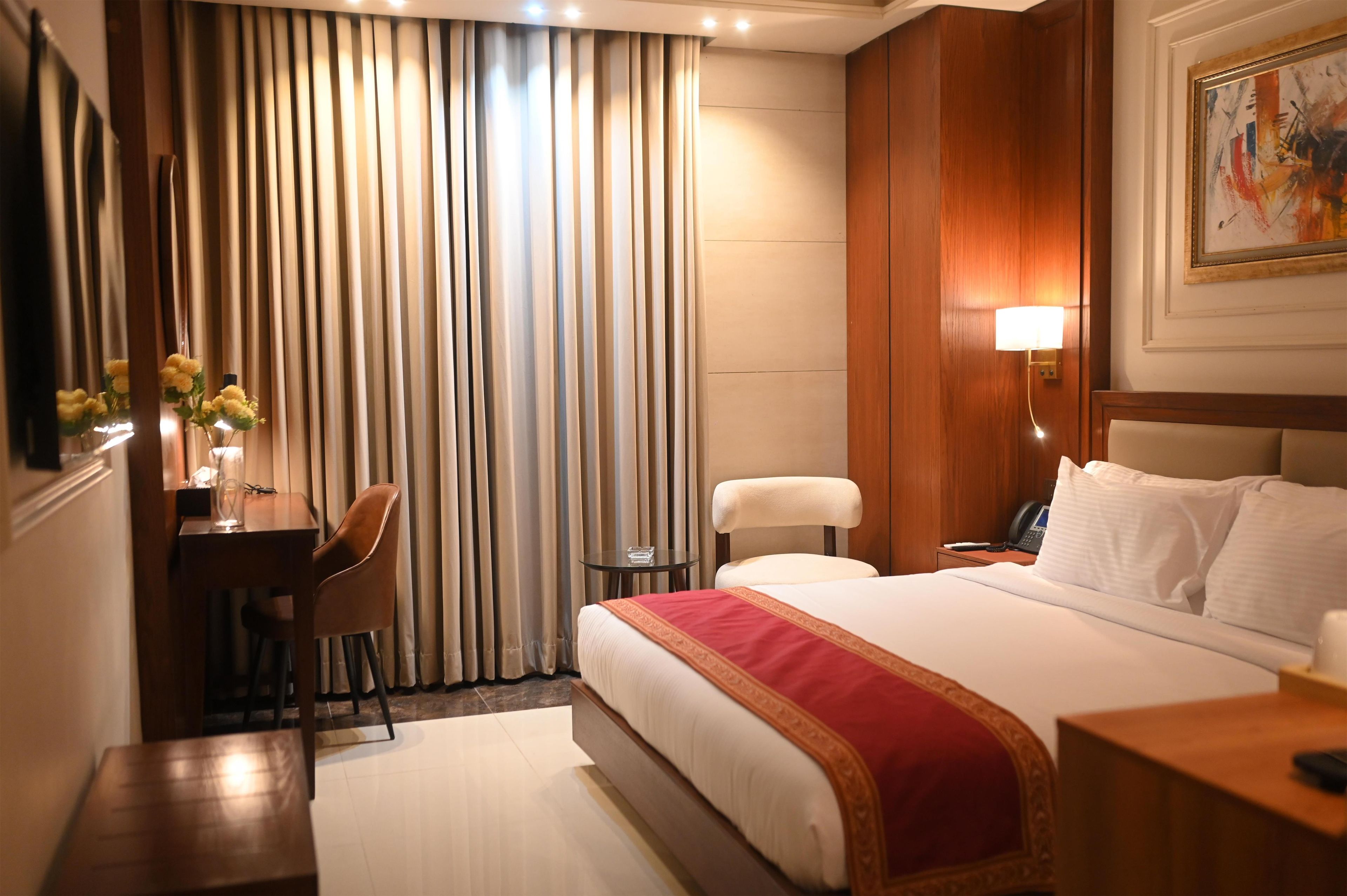 A hotel room with a bed, a desk with a chair, a bedside table with a lamp, and a painting on the wall.