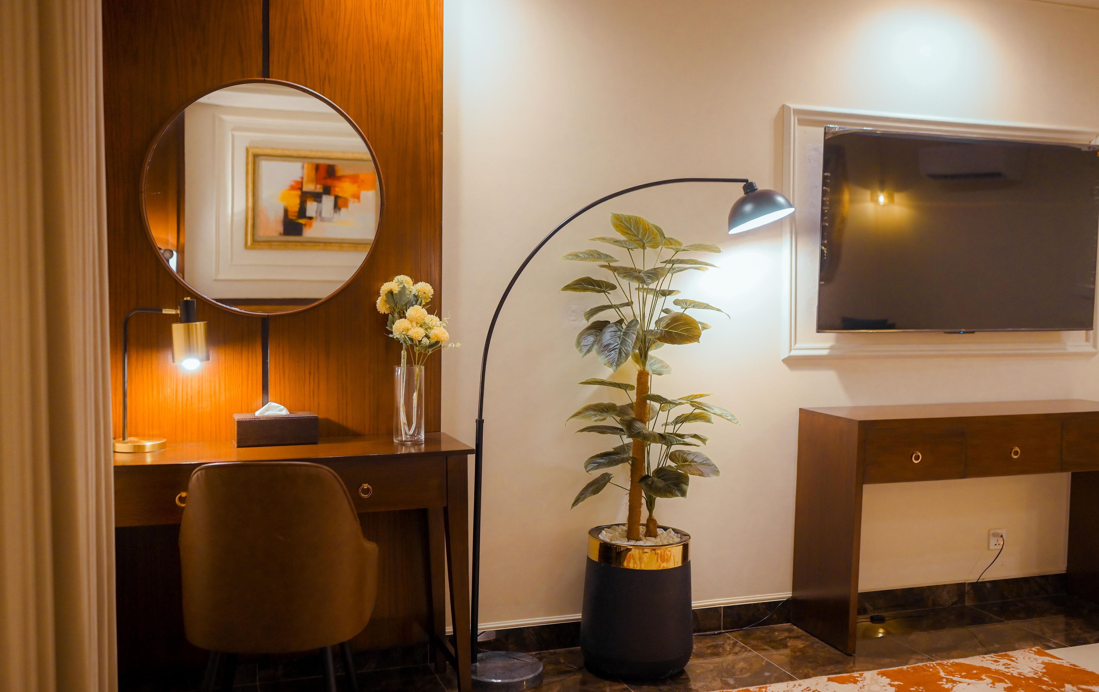 A hotel room with a desk, chair, mirror, lamp, potted plant, and a TV.