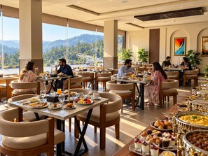 Un restaurante con varias mesas y sillas, un bufé con diversos alimentos, y vista al exterior.