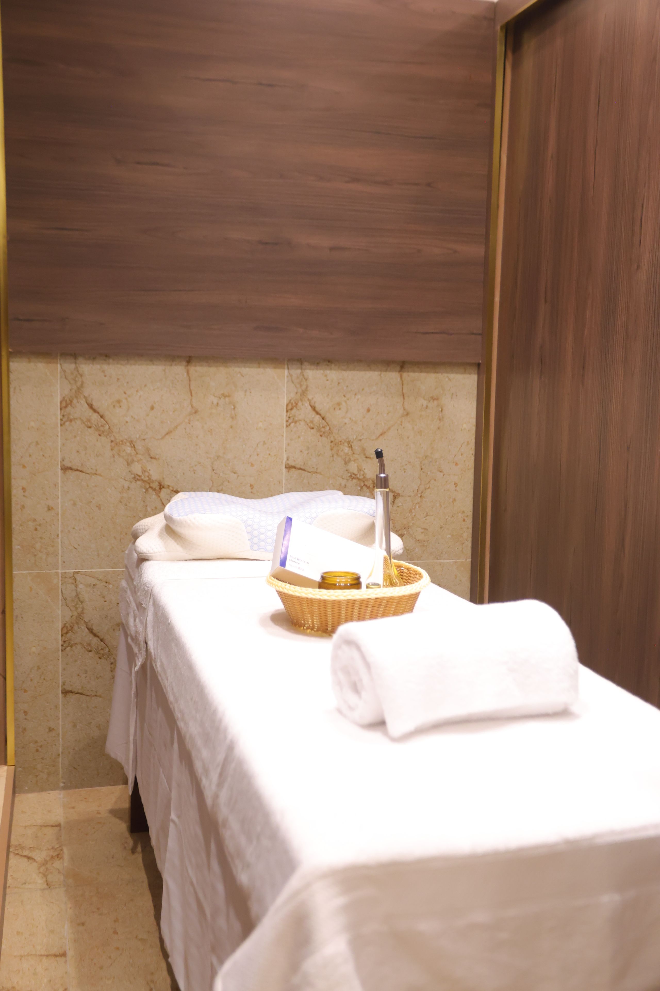 A neatly made bed with a white towel, a small basket, and a bottle, likely containing lotion or shampoo, in a room with a marble wall and wooden paneling.