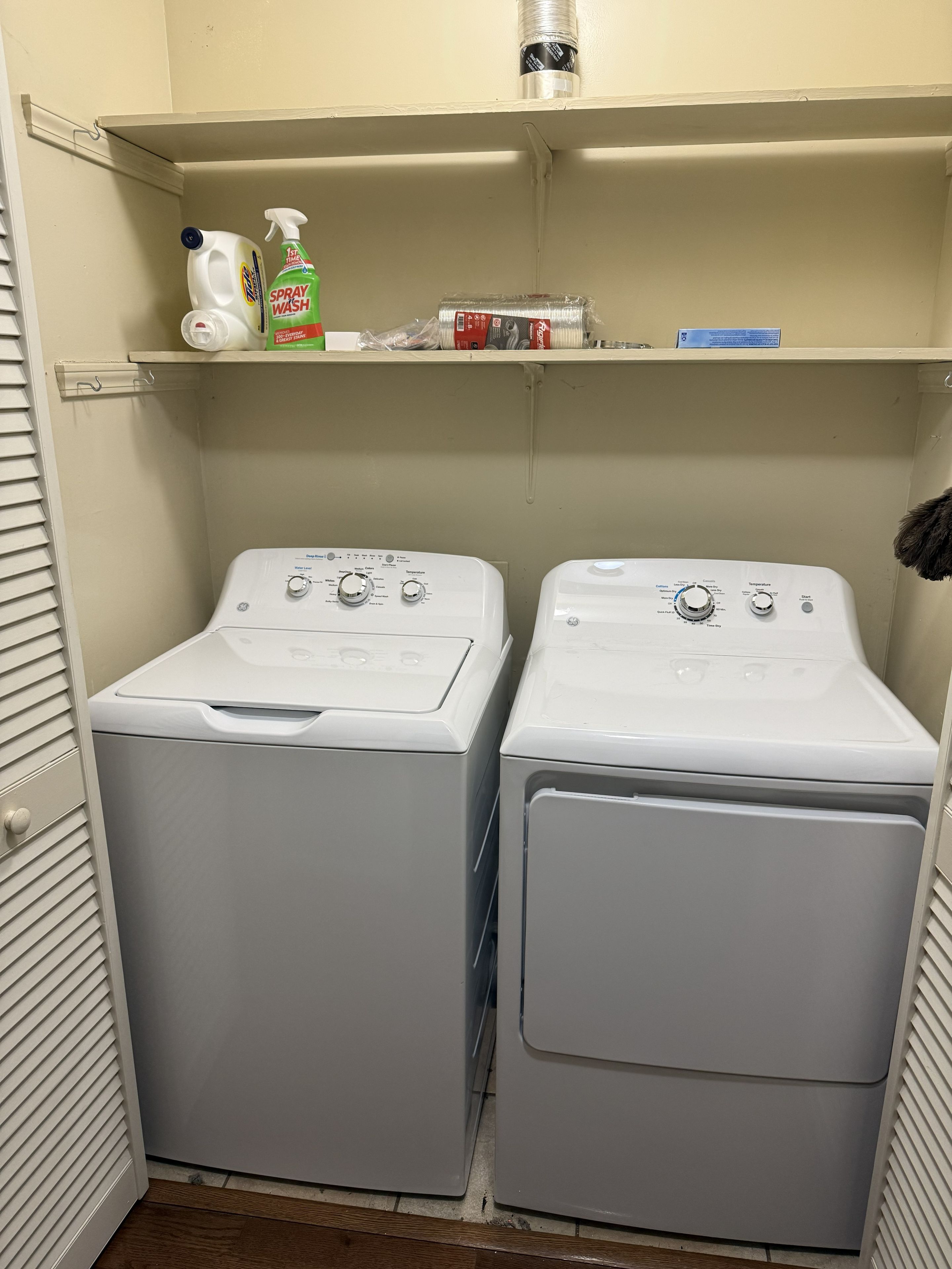 A laundry room with two front-loading washing machines and a shelf above containing cleaning supplies.