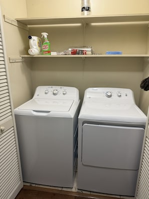 A laundry room with two front-loading washing machines and a shelf above containing cleaning supplies.