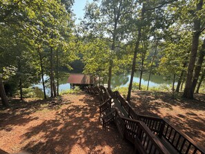 Une terrasse en bois mène à un chalet rustique situé au bord d’un lac, lui-même entouré d’arbres.