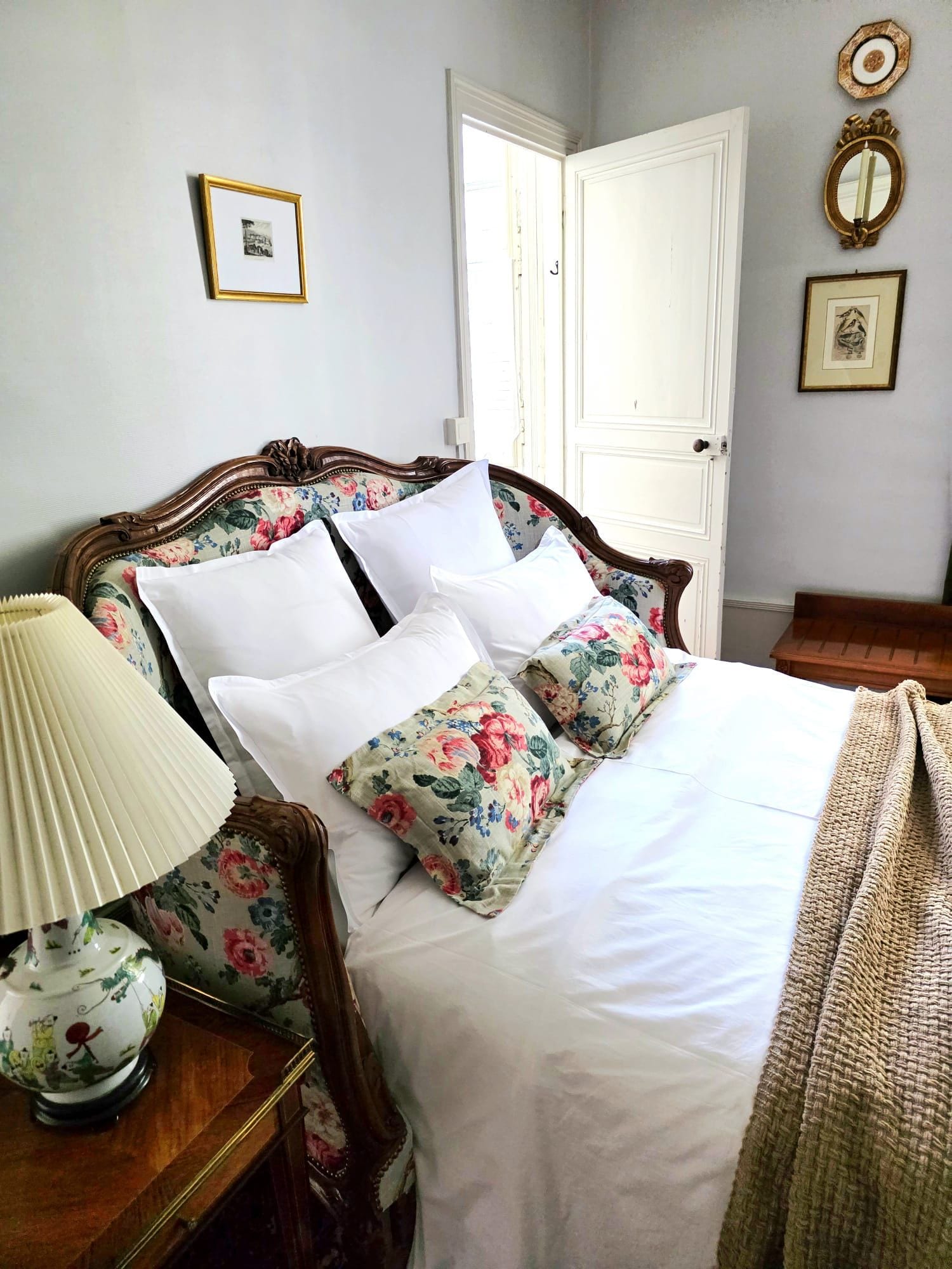 A neatly made bed with floral-patterned pillows and a white bedspread, a bedside table with a lamp, and a door with a mirror above it.