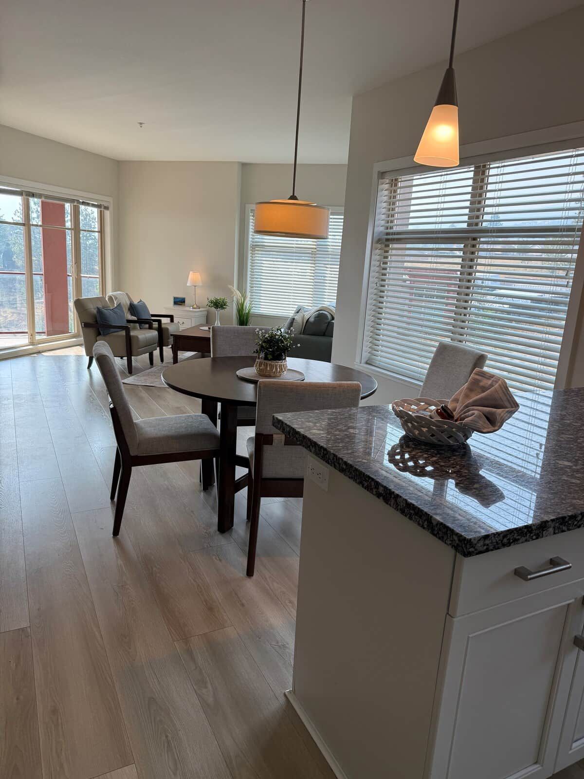 A modern kitchen with a granite countertop, a dining area with a table and chairs, and a large window with blinds.