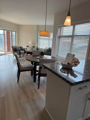 A modern kitchen with a granite countertop, a dining area with a table and chairs, and a large window with blinds. - Copper Sky Retreat (Central Okanagan)