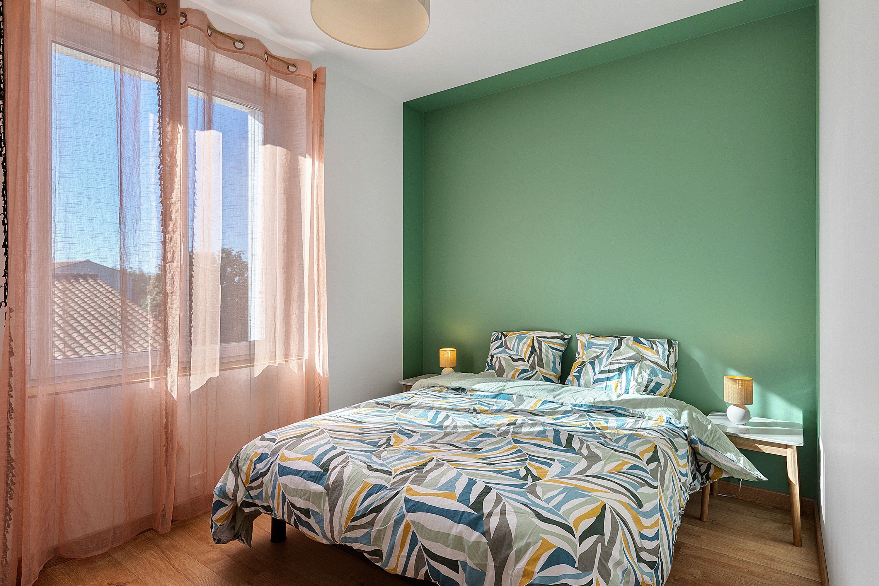 Une chambre à coucher avec un lit, une table de chevet, une lampe, des rideaux légers et une vue sur l’extérieur.