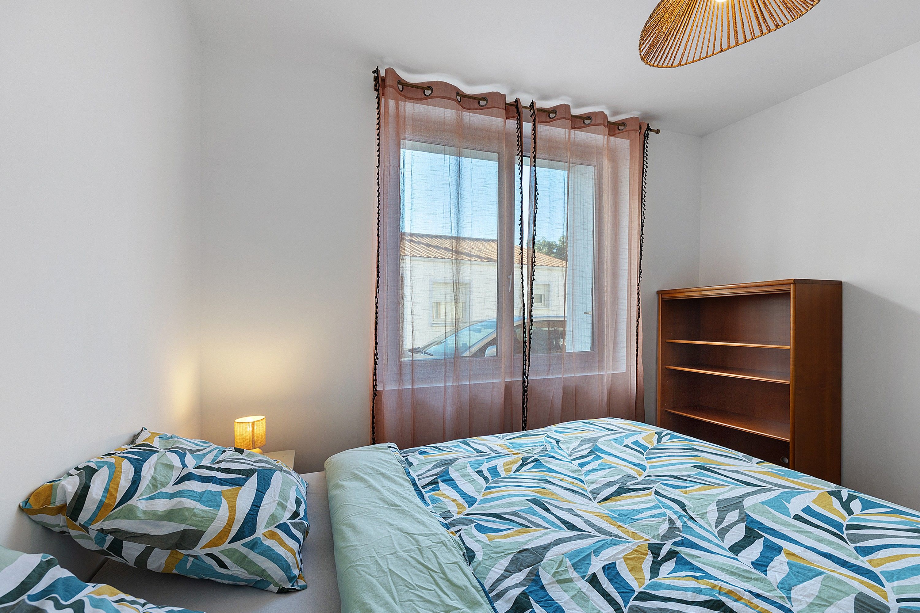 Une chambre à coucher avec un lit, une table de chevet avec une lampe, une fenêtre avec des rideaux et une bibliothèque en bois.