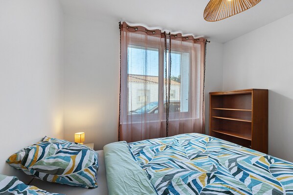 A bedroom with a bed, a bedside table with a lamp, a window with curtains, and a wooden bookshelf.