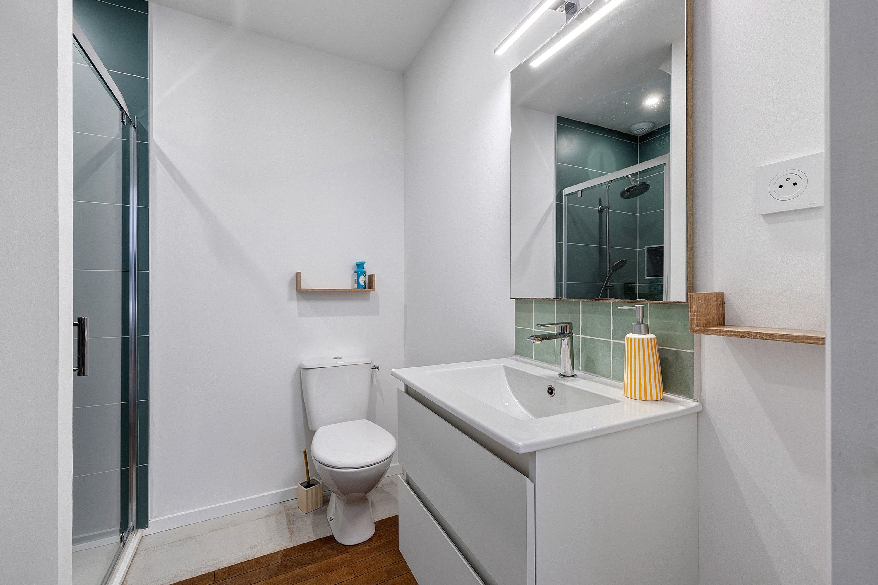 Une salle de bain moderne avec une douche à parois de verre, des toilettes blanches, un lavabo avec un miroir et une petite étagère contenant des articles de toilette.