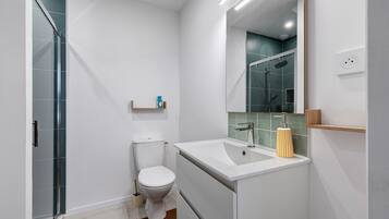 Une salle de bain moderne avec une douche à parois de verre, des toilettes blanches, un lavabo avec un miroir et une petite étagère contenant des articles de toilette.