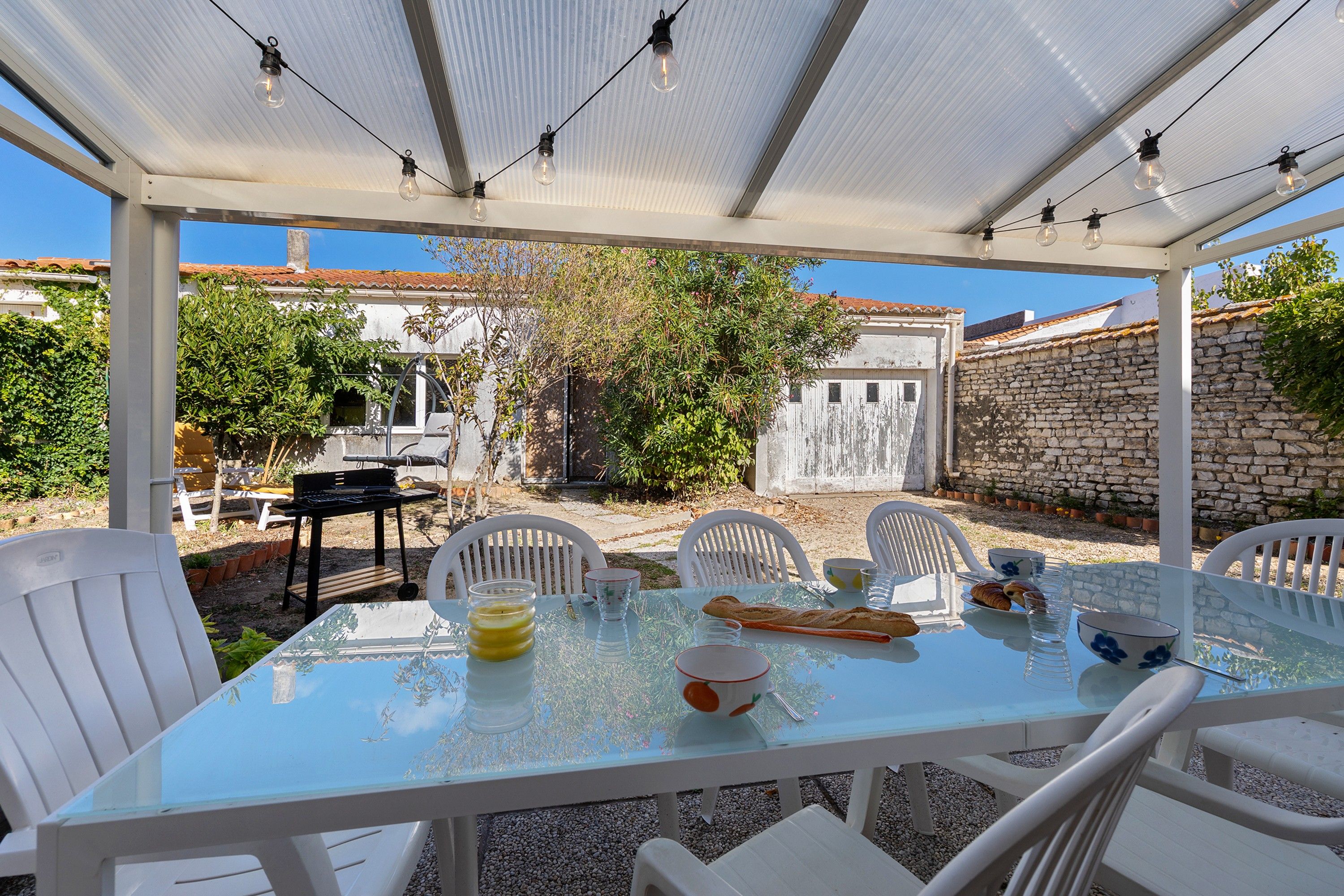Une terrasse extérieure avec une table en verre, des chaises blanches et des guirlandes lumineuses.