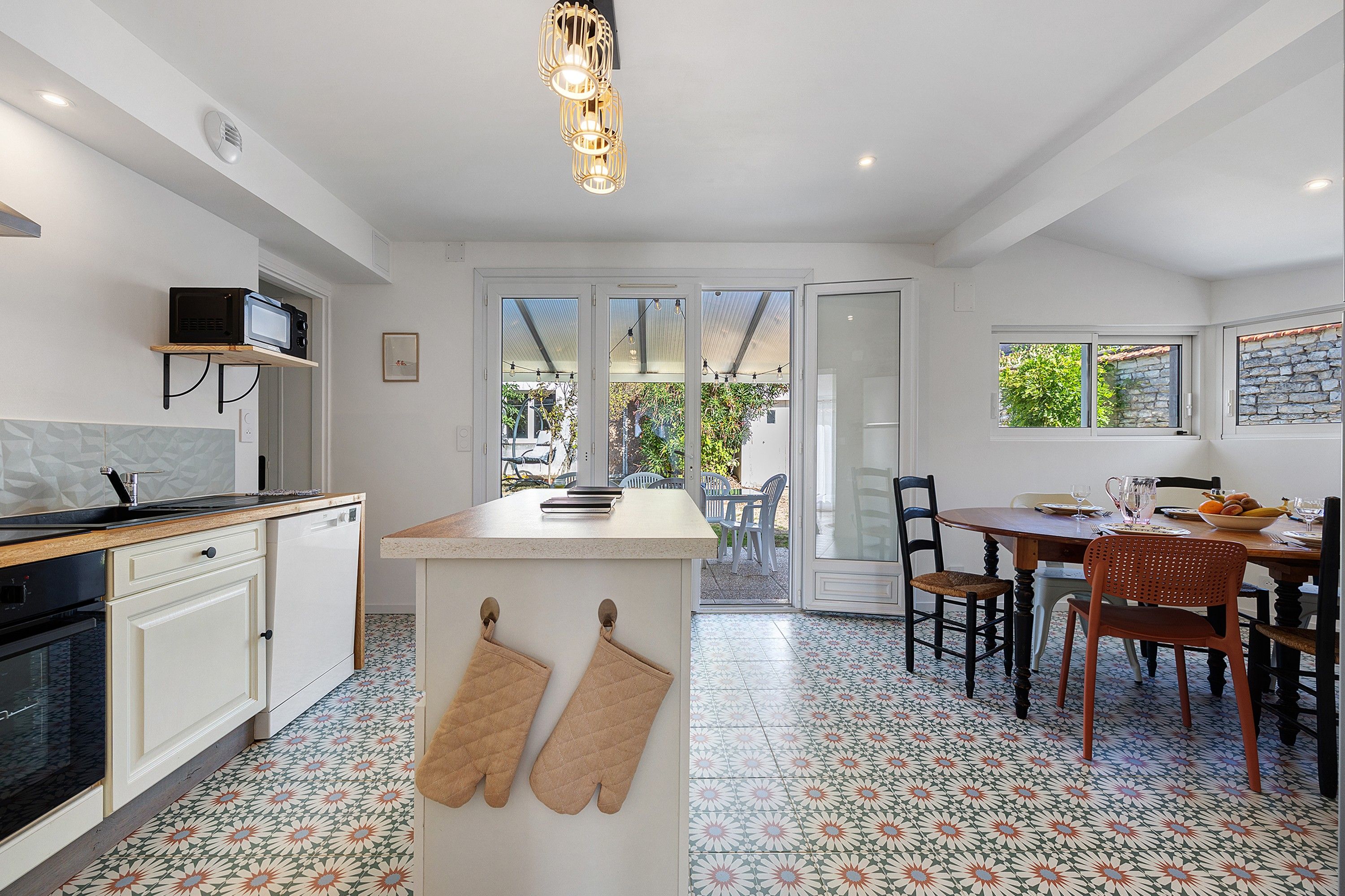 Une cuisine moderne avec un coin repas, comprenant un îlot central, un éclairage suspendu et une vue sur une terrasse extérieure à travers des portes vitrées.