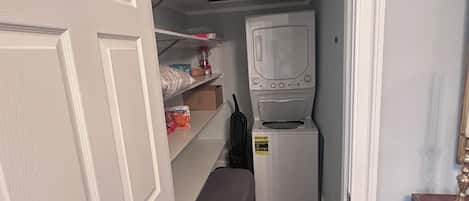 A narrow hallway with a white door on the left, a small laundry area with a washing machine and shelves above, and a dark wooden floor.