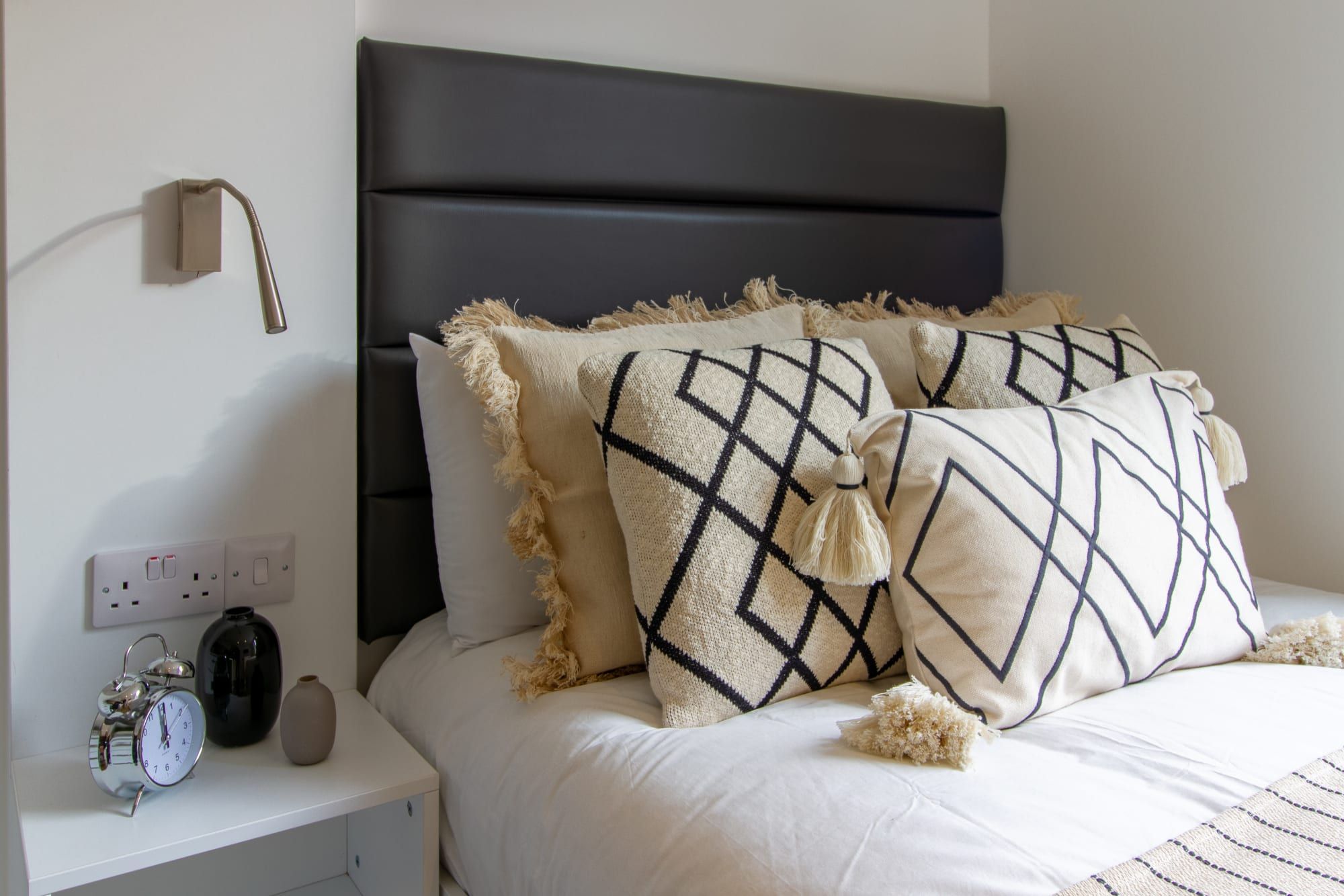 Un lit bien fait, avec une tête de lit, deux coussins décoratifs, une table de chevet avec une horloge et un vase, et une prise électrique au mur.