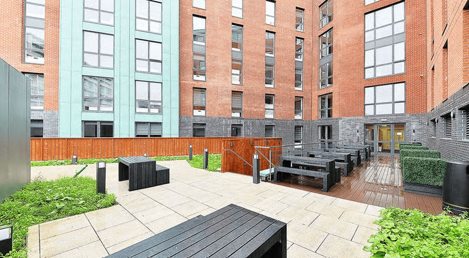 Un patio al aire libre con terraza de madera, bancos y un edificio de ladrillo con grandes ventanales.
