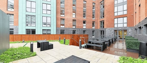 Un patio al aire libre con terraza de madera, bancos y un edificio de ladrillo con grandes ventanales.