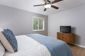 A bedroom with a bed, a television on a wooden cabinet, a ceiling fan, and a window with a view of trees. - Bradley Creek Retreat 5 min to Wrightsville Beach (Wilmington)