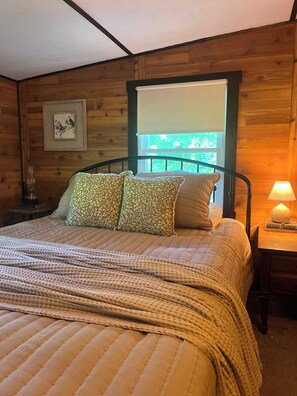 A wooden-paneled room with a bed, pillows, a nightstand, a lamp, and a window.
