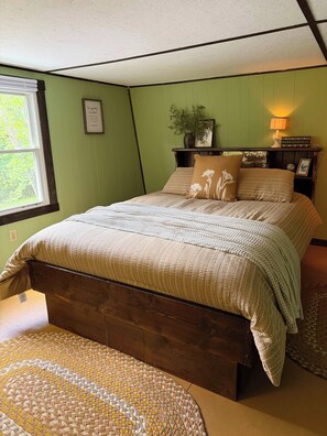 A bedroom with a wooden bed, a green wall, a window, and a bedside table with a lamp.