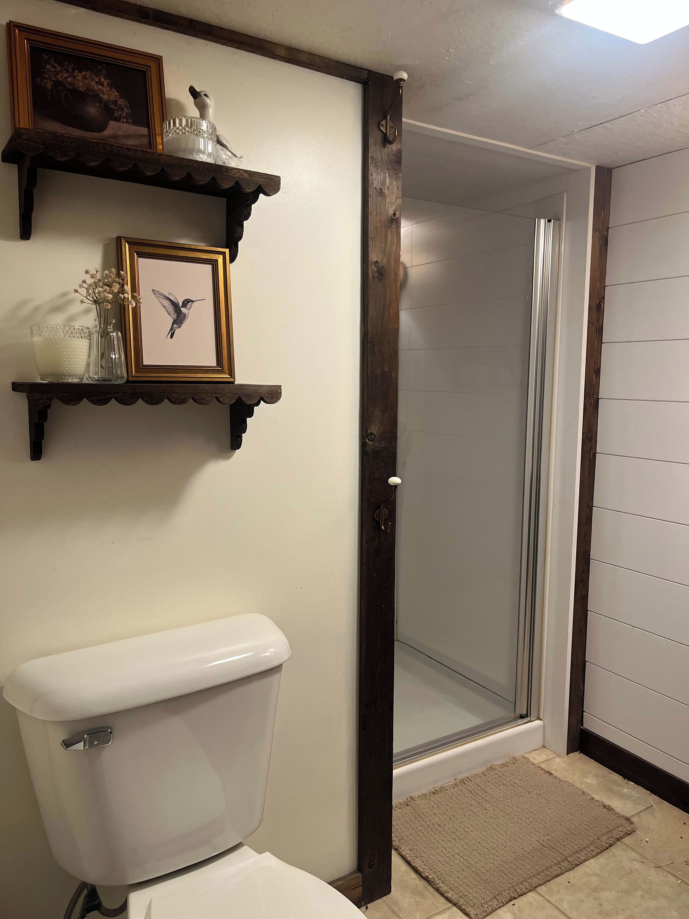 A bathroom with a toilet, a shower with a glass door, wooden shelves with decorative items, and a framed picture.