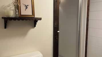 A bathroom with a toilet, a shower with a glass door, wooden shelves with decorative items, and a framed picture.
