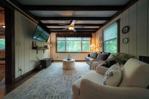 A living room with a ceiling fan, a flat-screen TV mounted on the wall, a sofa, a coffee table, and a window with blinds.