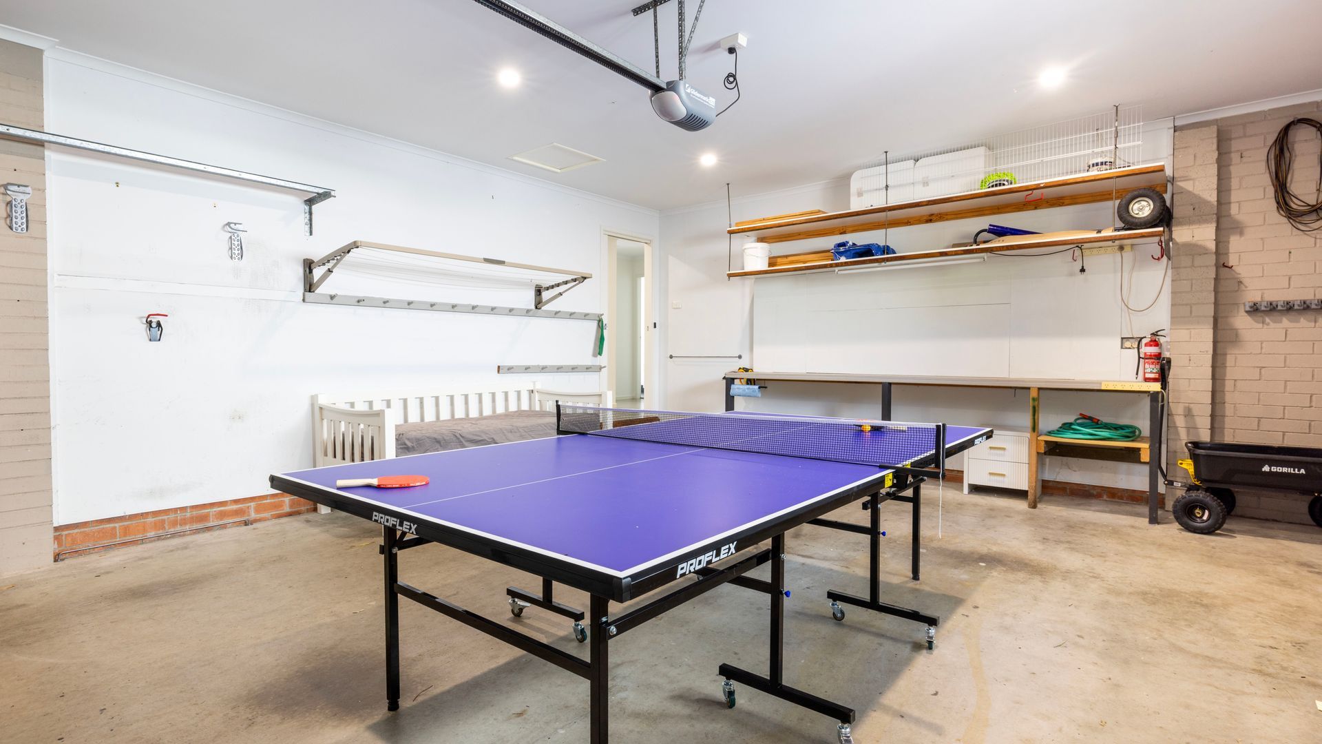 Una mesa de ping pong en una habitación con una puerta blanca, un radiador blanco y una estantería con varios objetos.