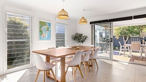 A dining area with a wooden table and white chairs, large windows, and a view of the outdoors. - Saltwood - Pet Friendly - Community Pool (Callala Beach)