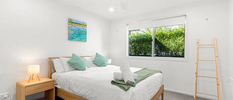 Un dormitorio con cama, mesita de noche, lámpara, ventana con vista a follaje y una escalera.