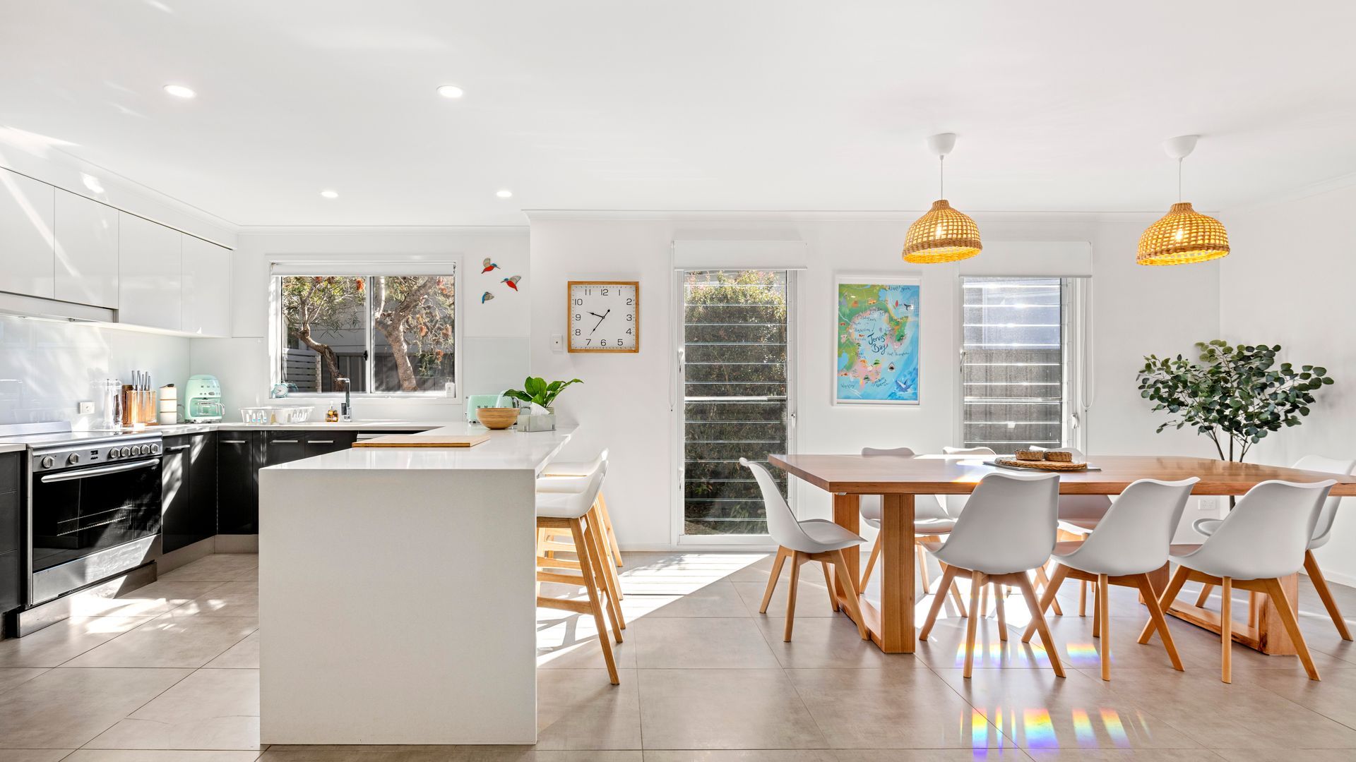 A modern kitchen with a central island, a dining area with a wooden table and white chairs, and a view of the outdoors through large windows.