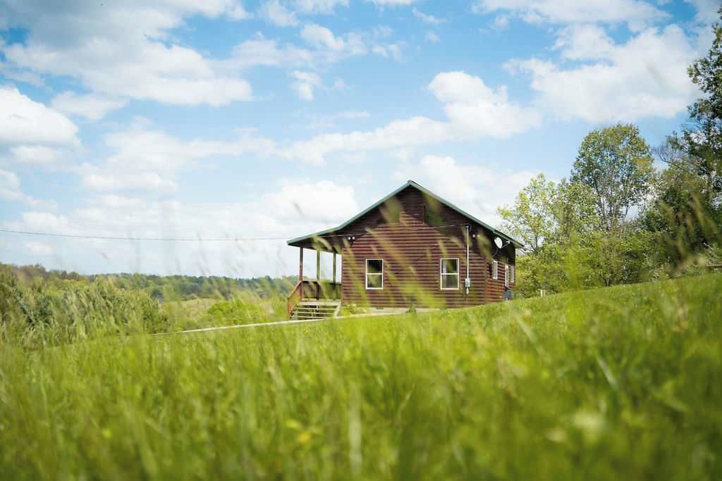 Et træhus med en veranda, omgivet af frodig græs og træer under en delvist skyet himmel.